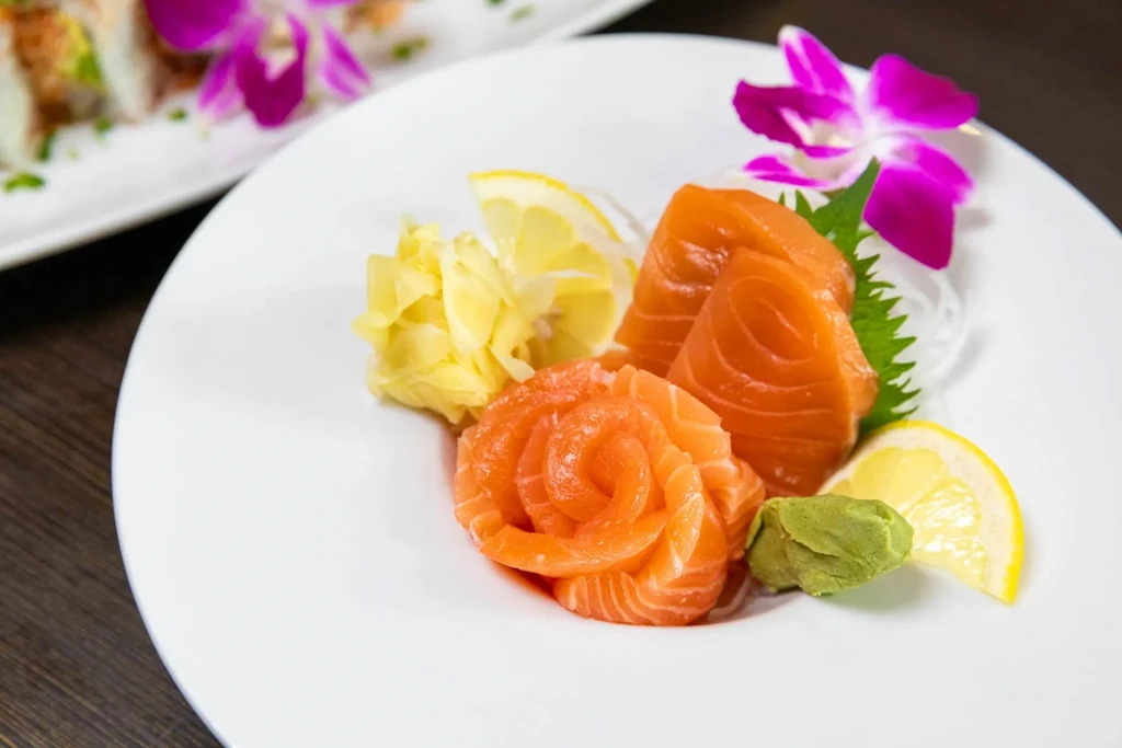 Close-up of salmon sashimi, ginger, wasabi, and lemon on a white plate.