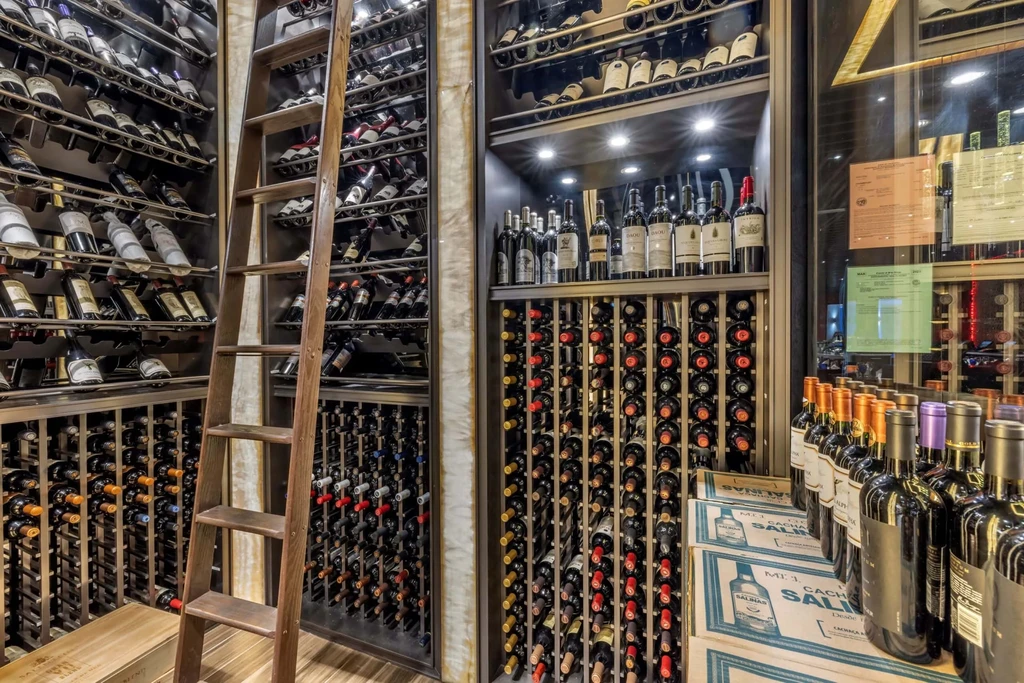 Well-stocked wine cellar with floor-to-ceiling bottles, ladder, and Cachaça boxes.