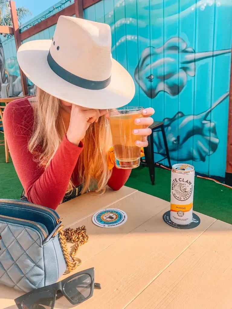 Person in hat sips drink at table with White Claw and stingray mural.