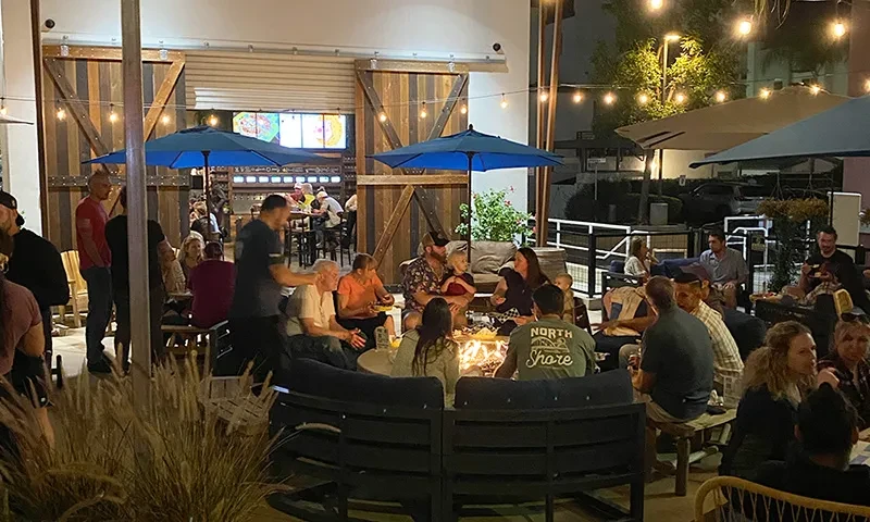 Evening restaurant patio with people, fire pit, string lights.