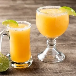 Two yellow-orange frozen drinks, rimmed with salt and lime, on wood.
