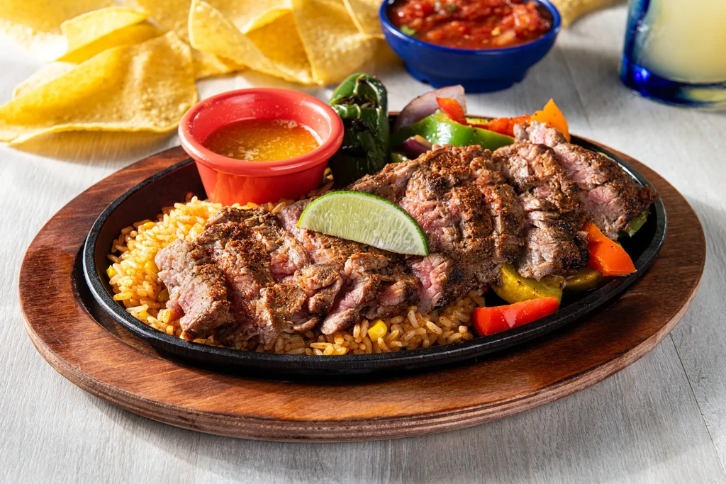 Cast-iron platter of sizzling steak, rice, grilled peppers, and onions.