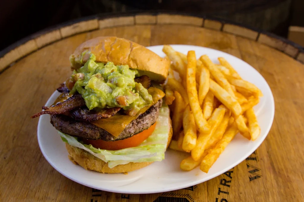 Hamburger with cheese, bacon, guacamole, and fries on a white plate.