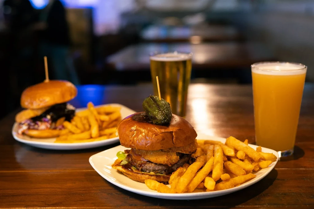 Two plates of burgers, fries, and beers on a dark table.