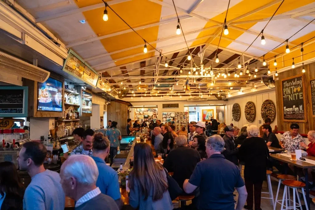 Crowded bar interior, patrons under string lights and striped ceiling.