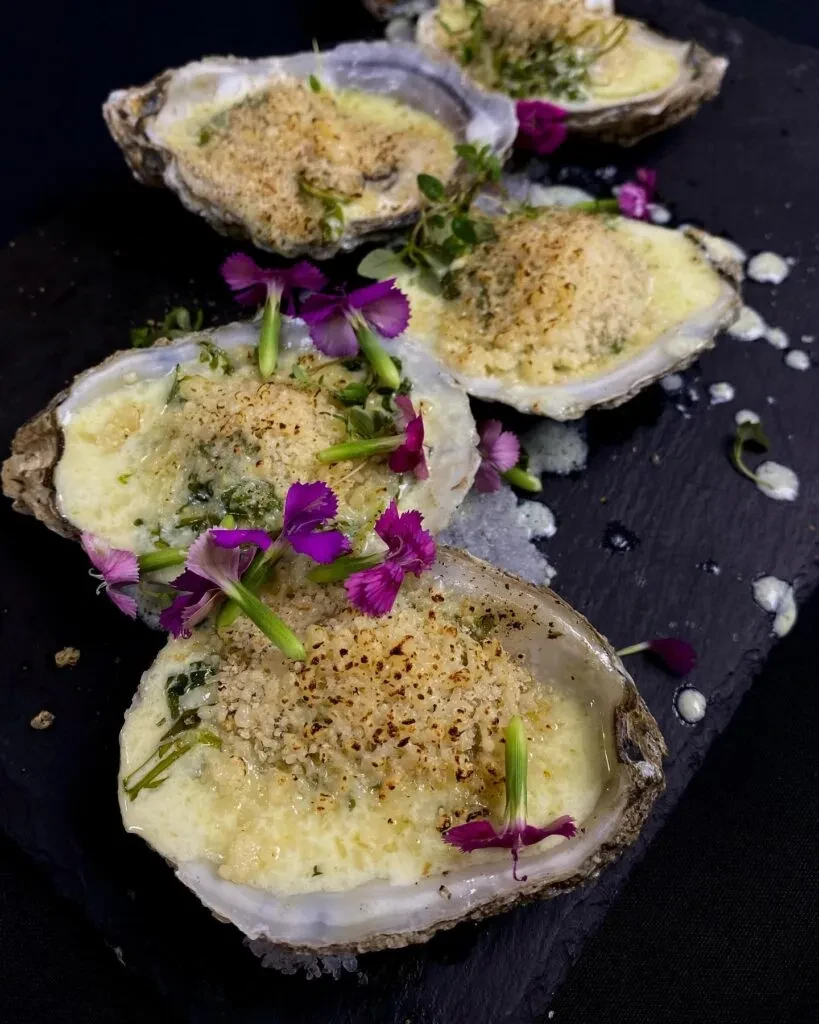Baked oysters topped with creamy sauce, breadcrumbs, herbs, and purple flowers.