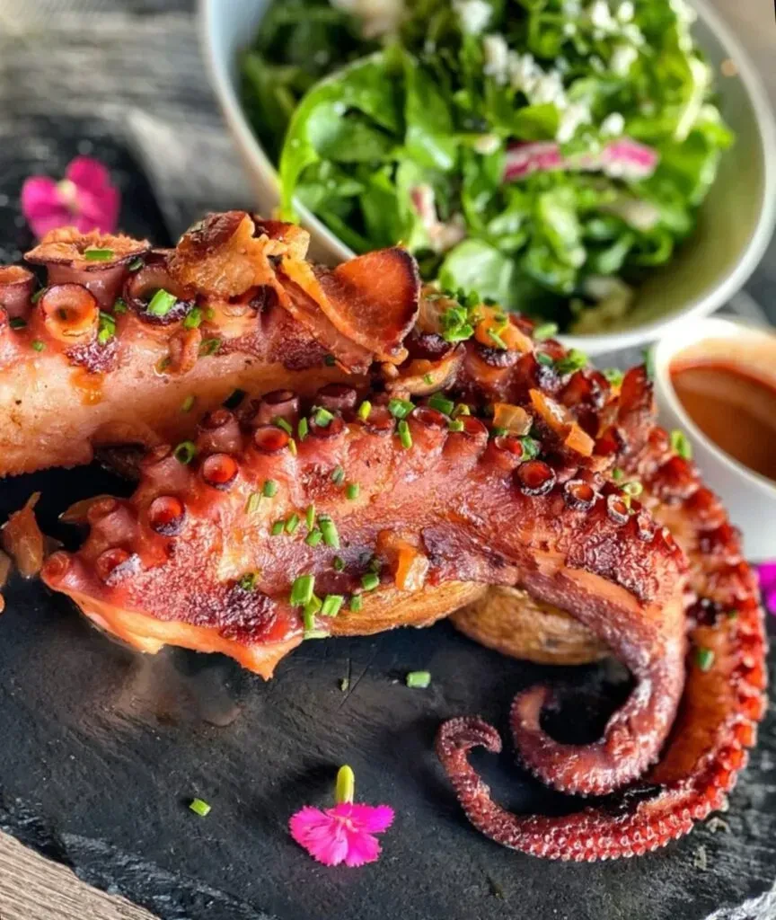 Charred grilled octopus, chives, flower on slate, with salad and sauce.