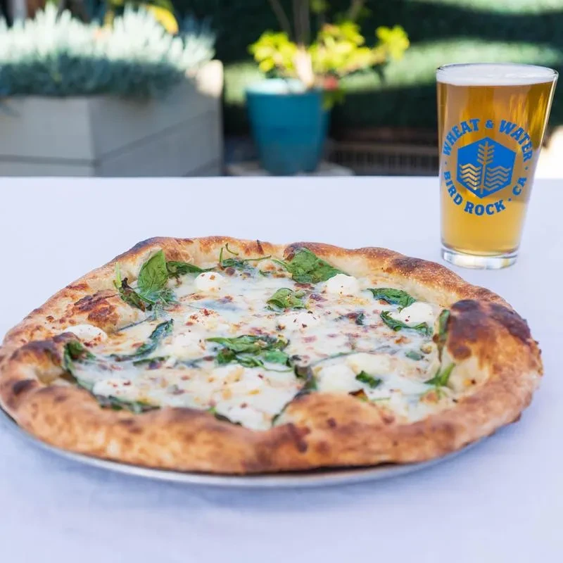 Close-up of spinach pizza, beer, and white tablecloth outdoors.