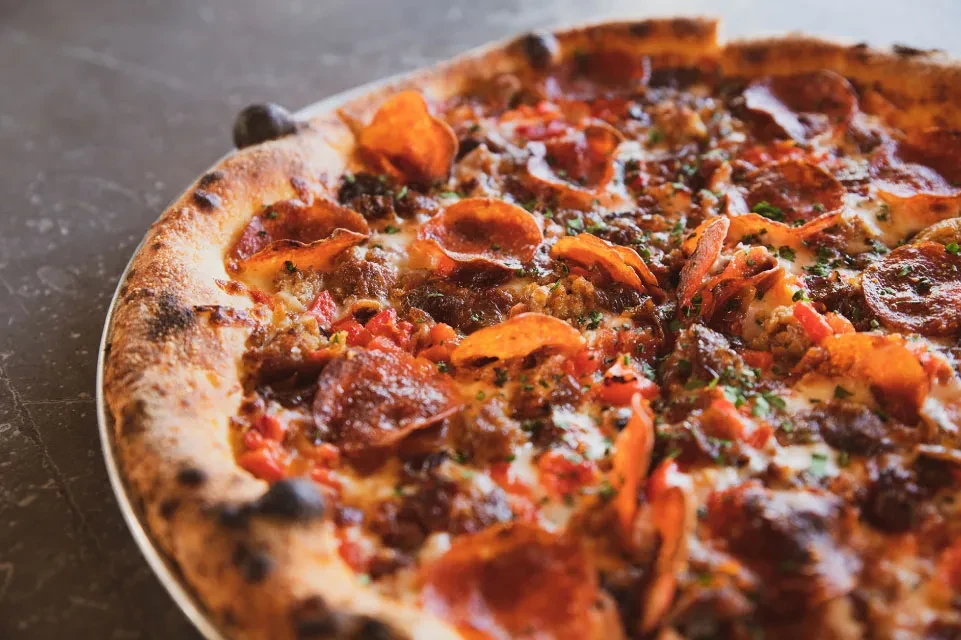 Close-up of wood-fired pizza with pepperoni, sausage, peppers, cheese, and herbs.