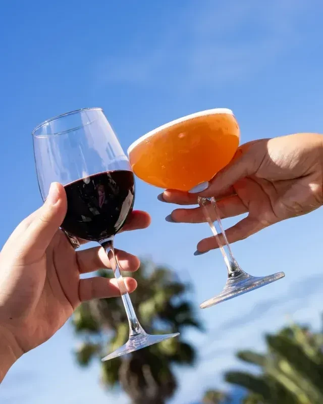 Hands toast red wine and frothy cocktail against blue sky.