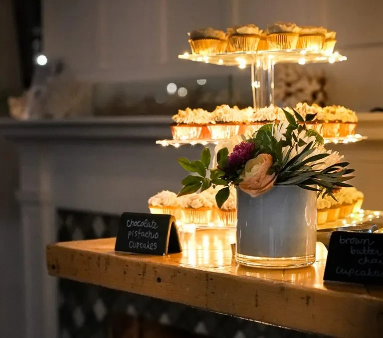 Tiered cupcake display with fairy lights and flavor signs on a rustic counter.