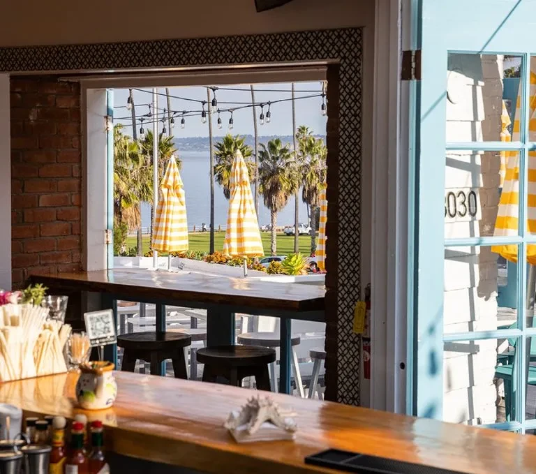 View from a bar overlooking ocean, palm trees, and striped umbrellas.