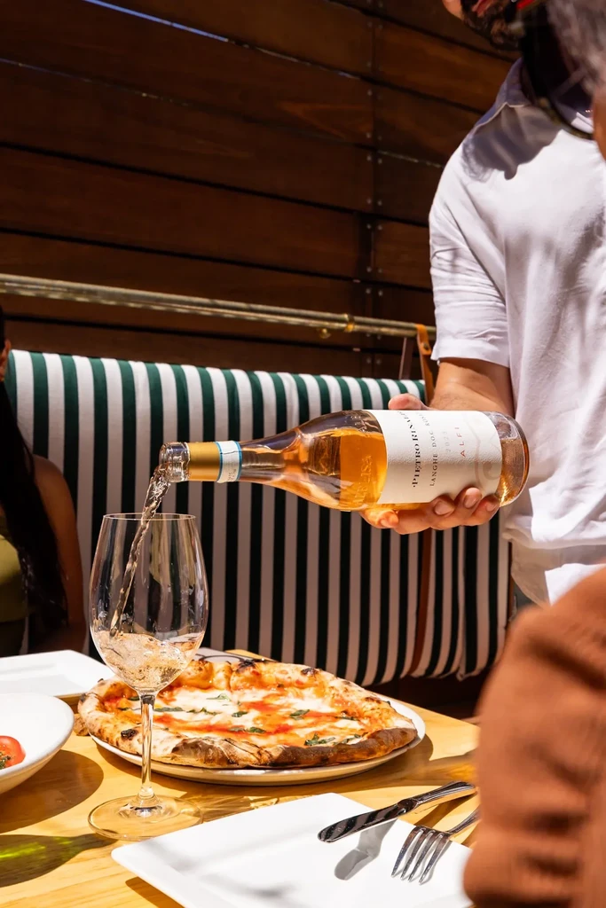 Hand pouring rosé wine into glass, pizza on sunlit table.