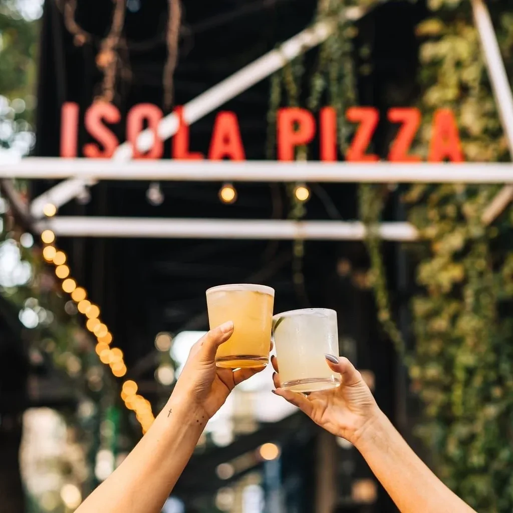 Hands toast amber and lime drinks at Isola Pizza with string lights.