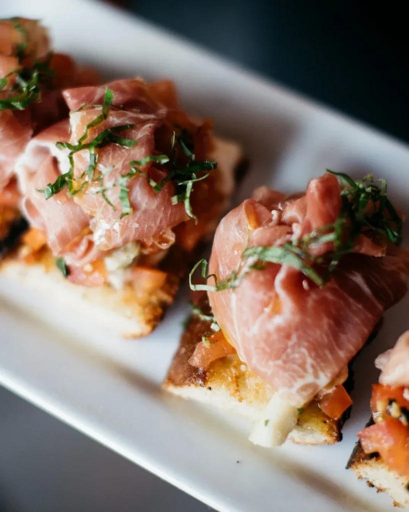 Close-up of crostini topped with prosciutto, tomatoes, and fresh green herbs.