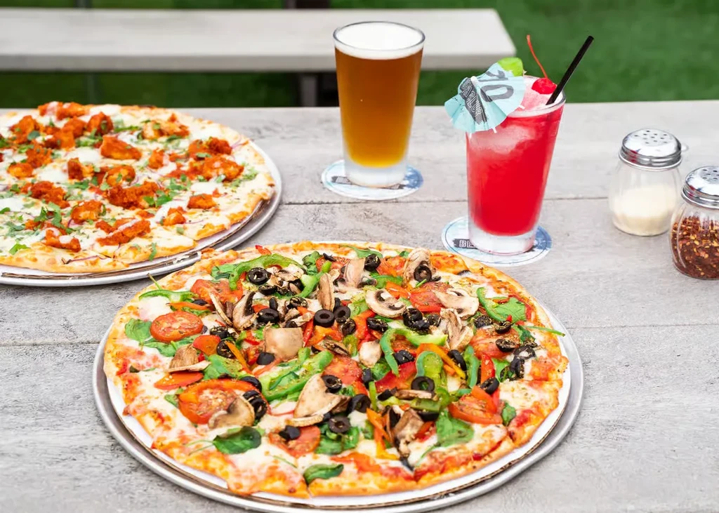Outdoor dining with two pizzas, two drinks, and seasoning shakers.