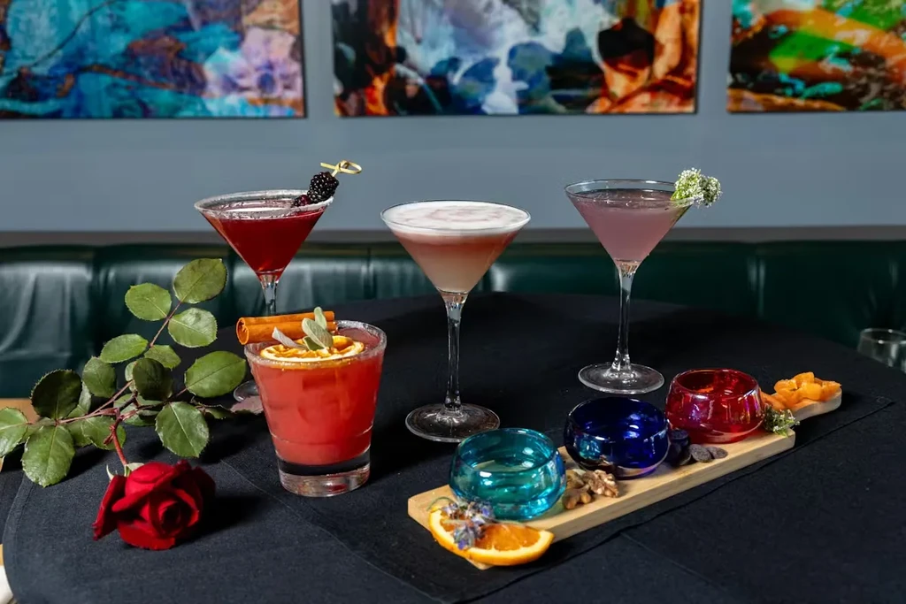 Four garnished cocktails, a rose, votives, and snacks on black table.