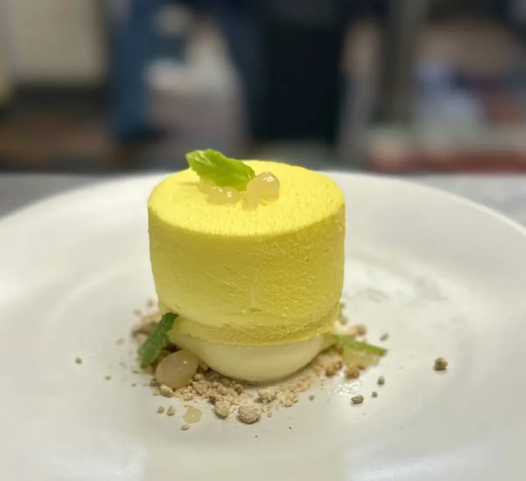 A beautifully plated yellow cylindrical dessert with mint and pearls on crumbs.