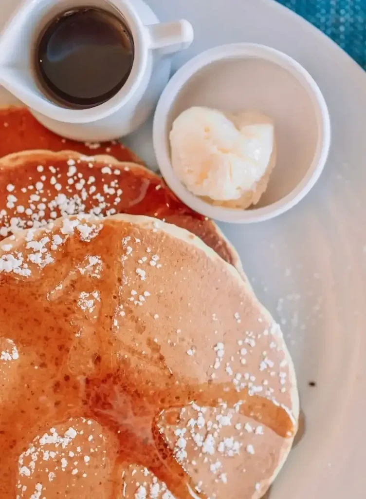 Stacked pancakes with syrup, powdered sugar, heart-shaped butter, and syrup pitcher.