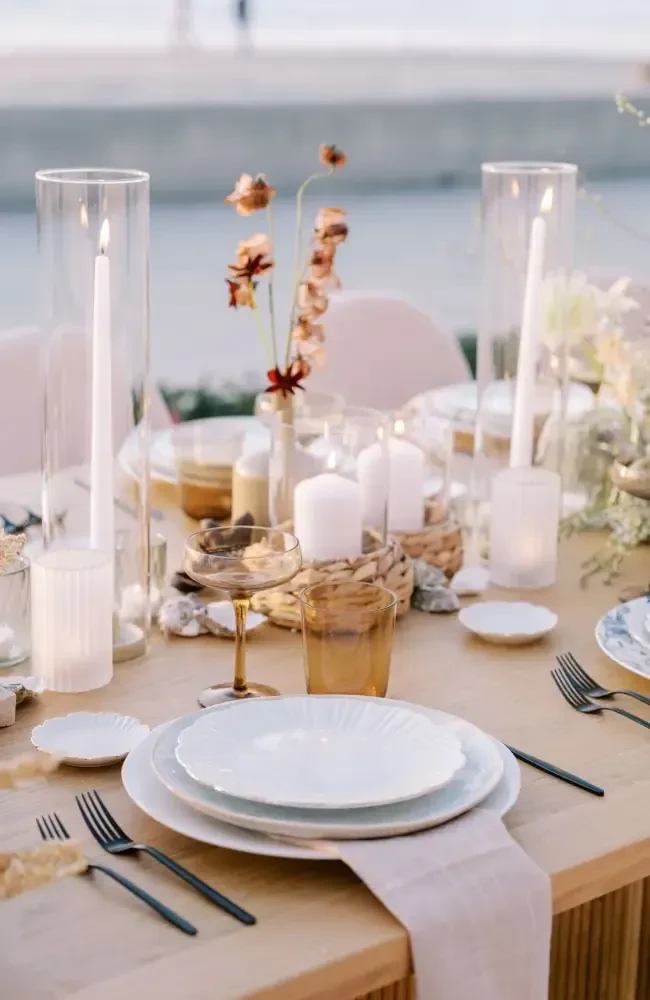 Set dining table with white plates, lit candles, rustic flowers, amber glasses.