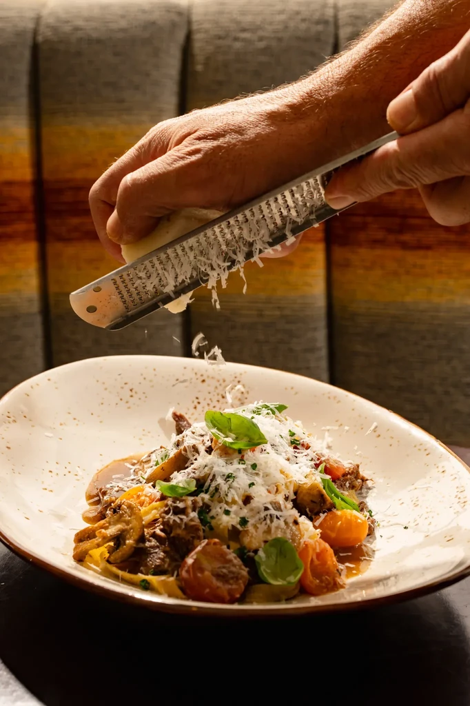 Hands grating hard cheese over a pasta and meat dish.