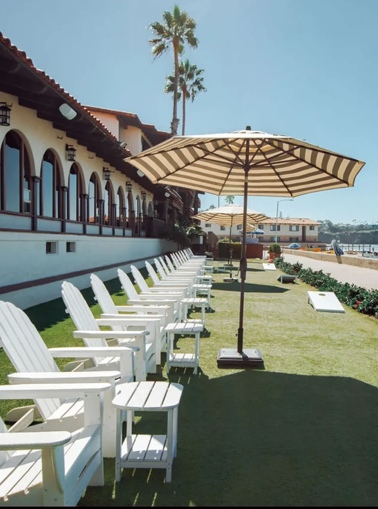 Outdoor amenity with white Adirondack chairs, striped umbrellas, Spanish architecture, ocean.