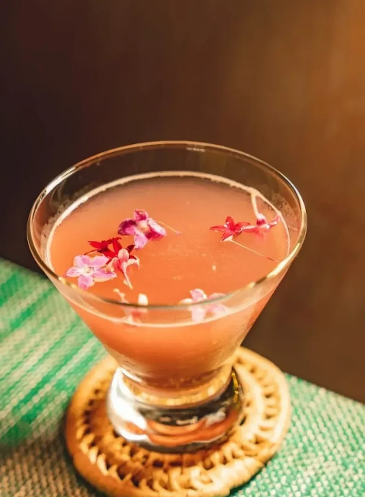Coral cocktail in a martini glass, garnished with purple and red flowers.