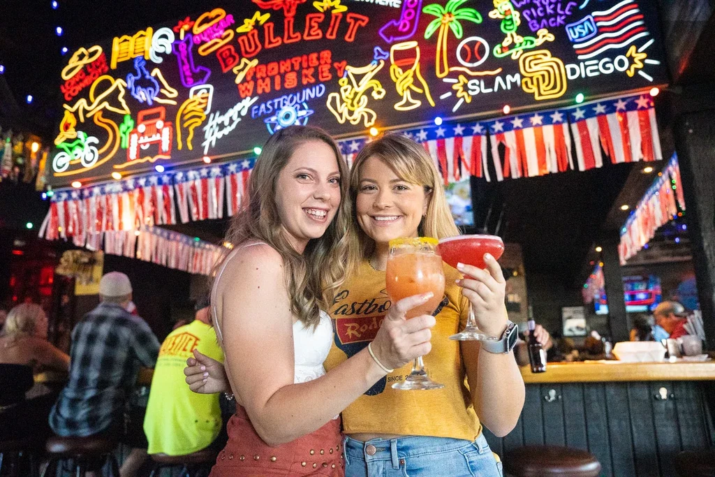 Two smiling women hold cocktails in a vibrant bar setting.
