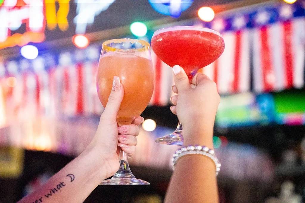 Two hands toast colorful cocktails with sparkling nails against a blurred background.