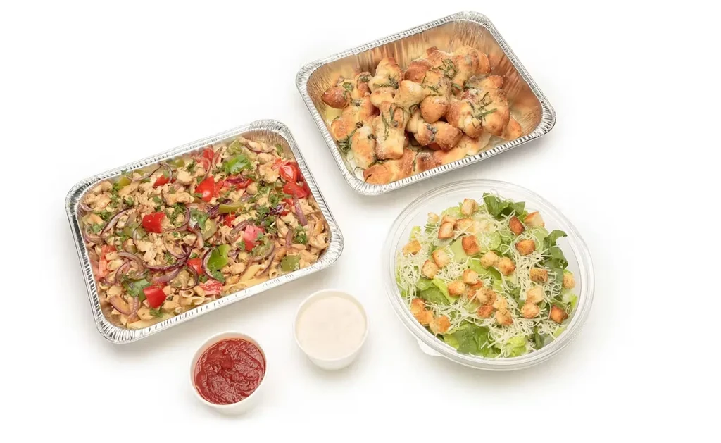 Flat lay of takeout chicken pasta, garlic knots, Caesar salad, sauces.