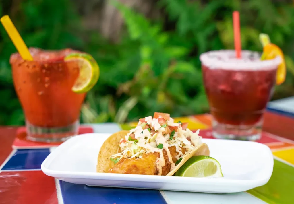 Single fried fish taco with toppings, two colorful drinks on a mosaic table.