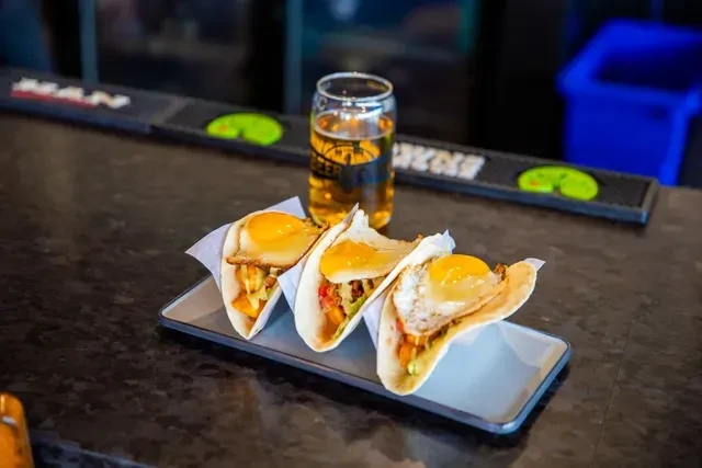 Three breakfast tacos with sunny-side eggs and beer on a bar.