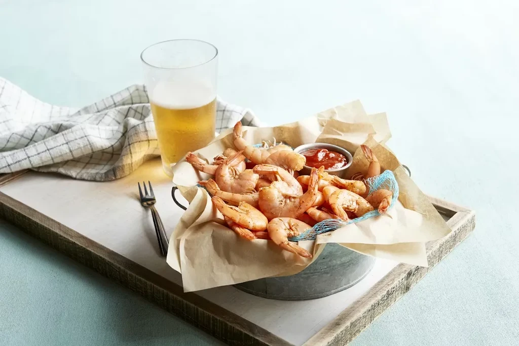 Shrimp bucket with red sauce, beer, and fork on a white tray.