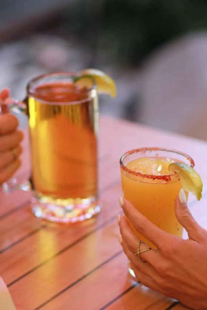 Two hands hold a chili-rimmed blended drink and a beer with lime.