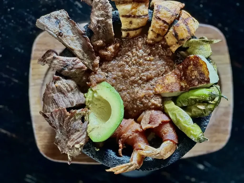 Overhead view of grilled molcajete with steak, chicken, shrimp in stone bowl.