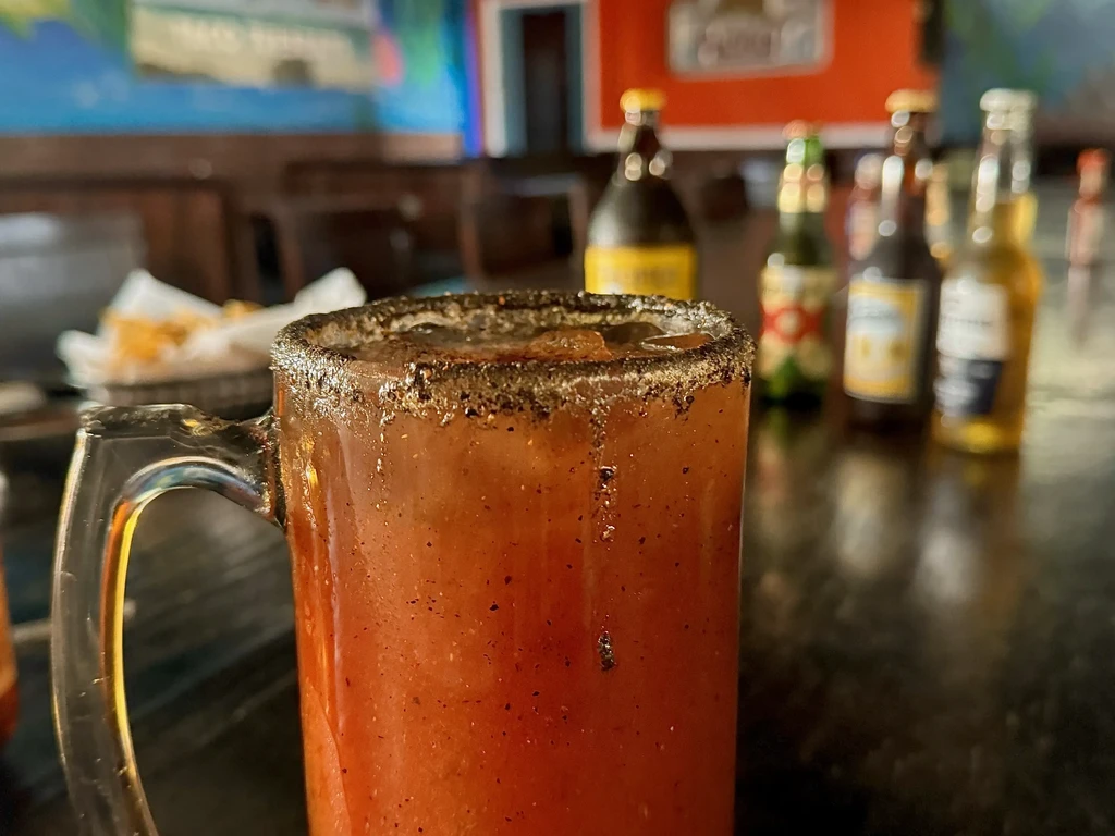Spice-rimmed reddish drink with blurred beer bottles, snacks, and mural background.