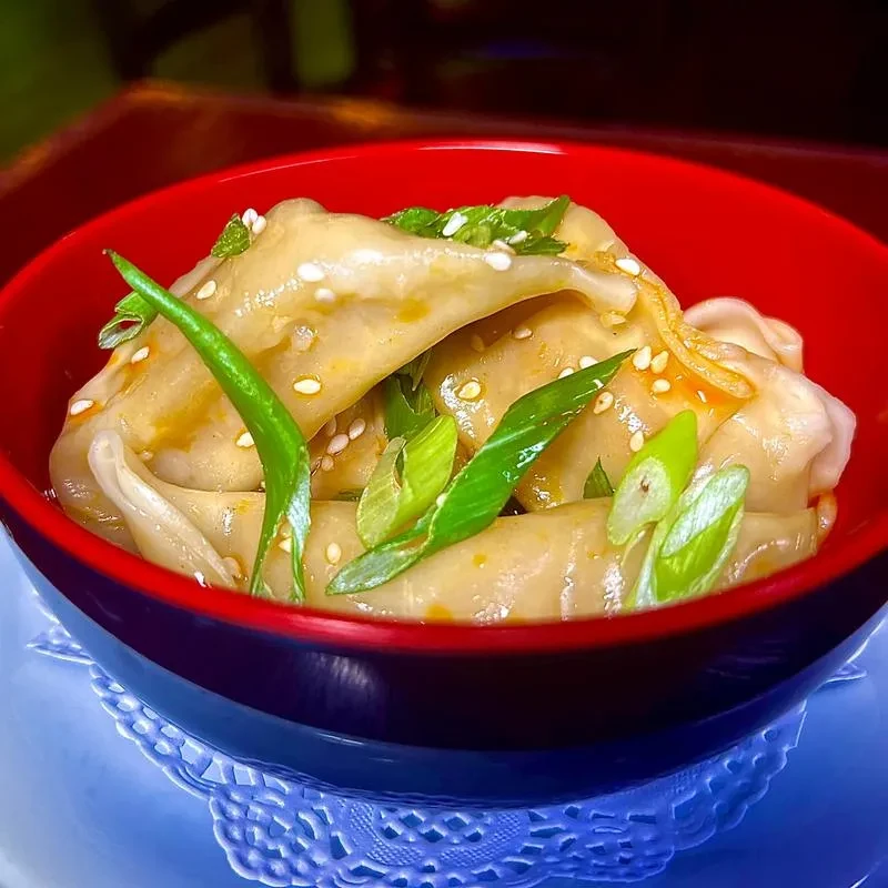 Dumplings in a red bowl with green onions, sesame seeds, and amber sauce.