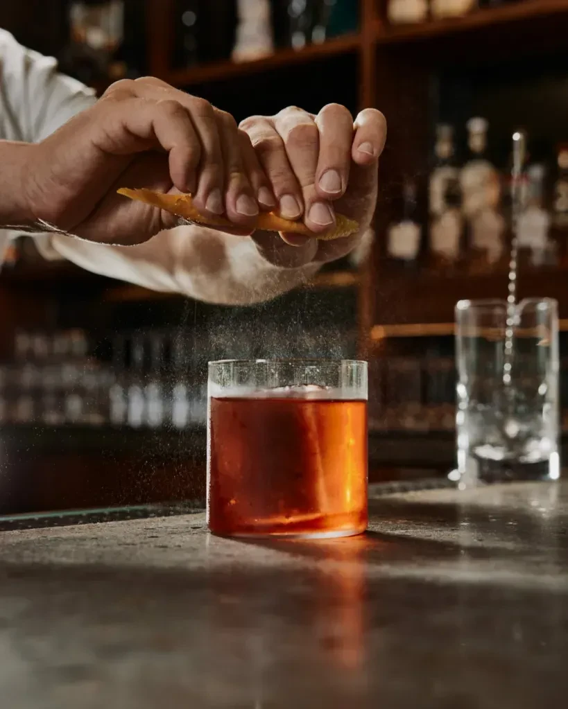 Close-up: hands express orange zest over a reddish-brown cocktail.