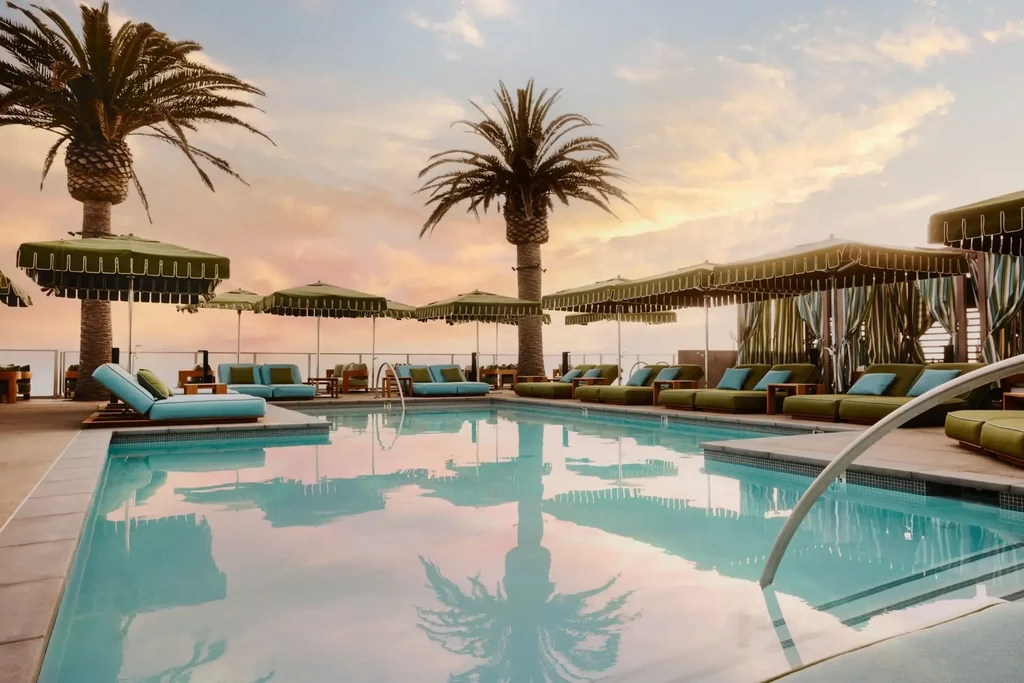 Rooftop pool area with lounge chairs, palm trees, reflecting colorful sky.