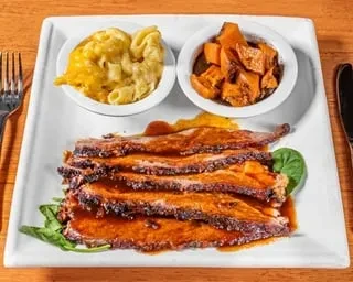 Sliced dark-glazed meat, mac and cheese, sweet potatoes on a plate.