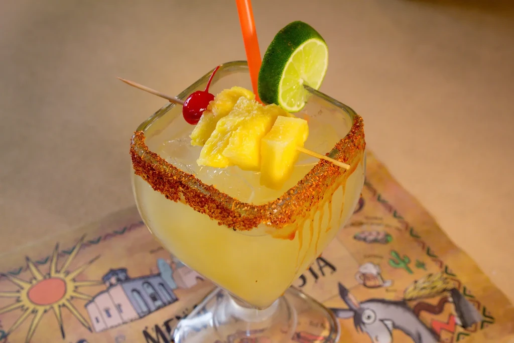 Margarita-style drink with spicy rim, lime, pineapple, and cherry skewer.