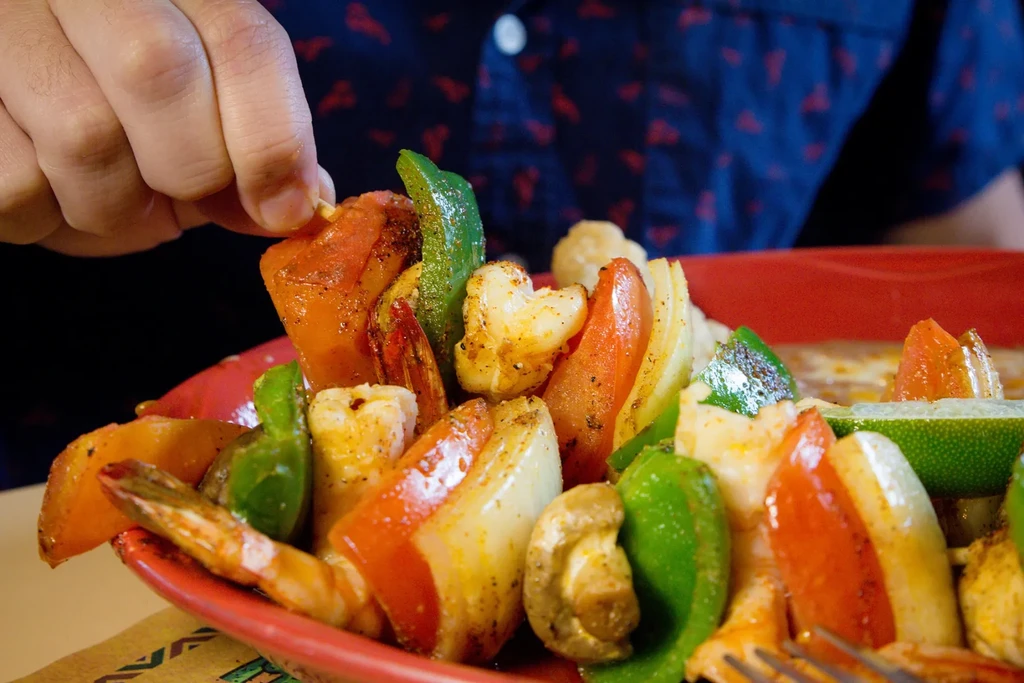 Hand holding colorful cooked shrimp and vegetable skewer on red plate.