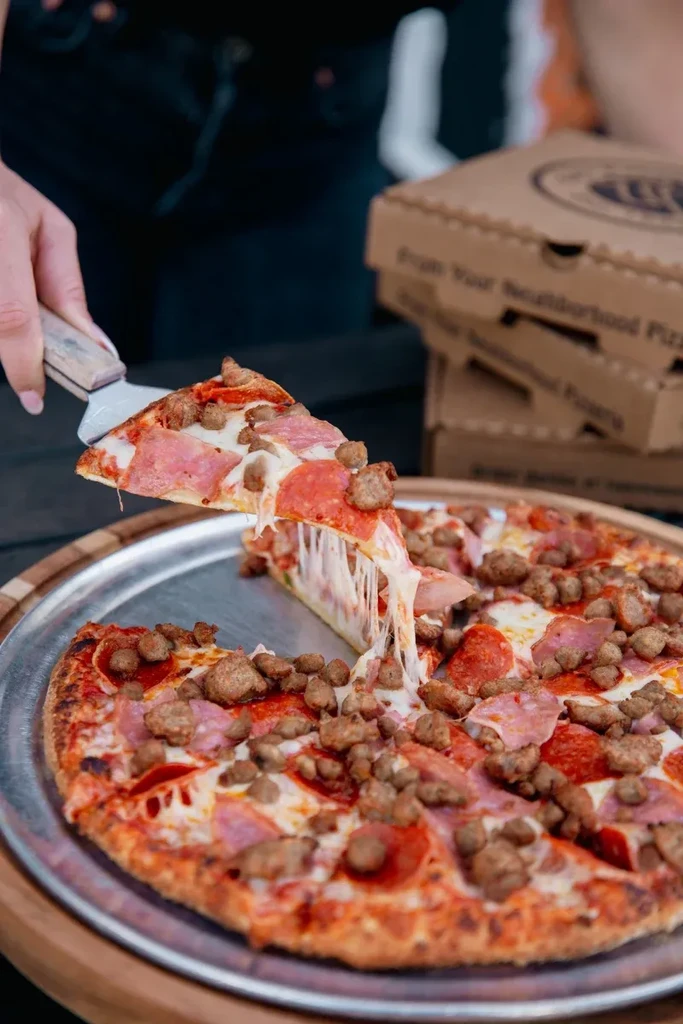 A hand lifts a slice of multi-meat pizza, cheese stretching.