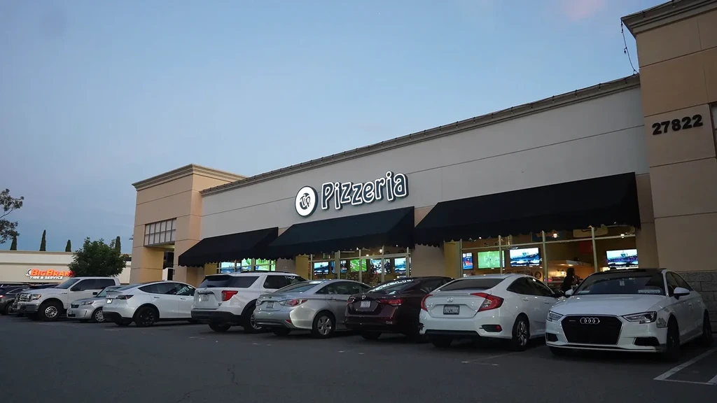 Illuminated Pizzeria storefront at dusk with cars in parking lot.