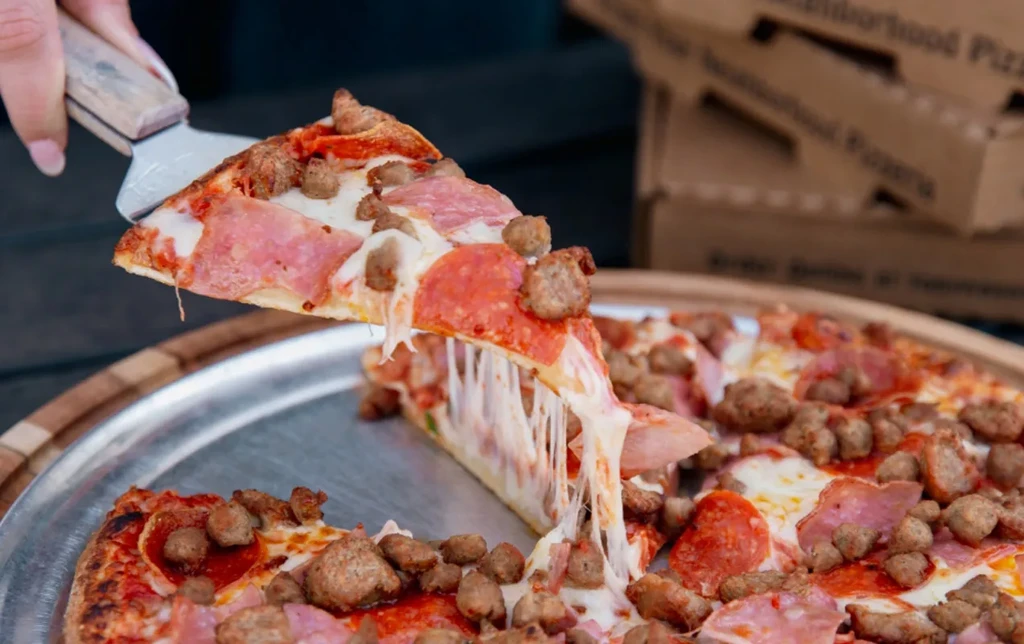 Hand lifting a cheesy slice of pepperoni, ham, and sausage pizza.