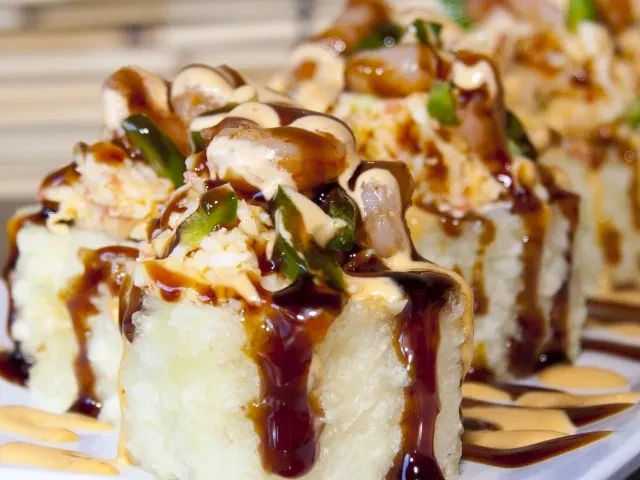 Close-up of crispy sushi rolls topped with seafood, greens, and two sauces.