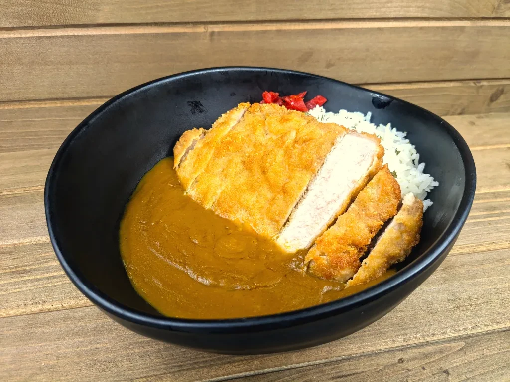 Sliced katsu curry with rice, sauce, and red pickles in a black bowl.