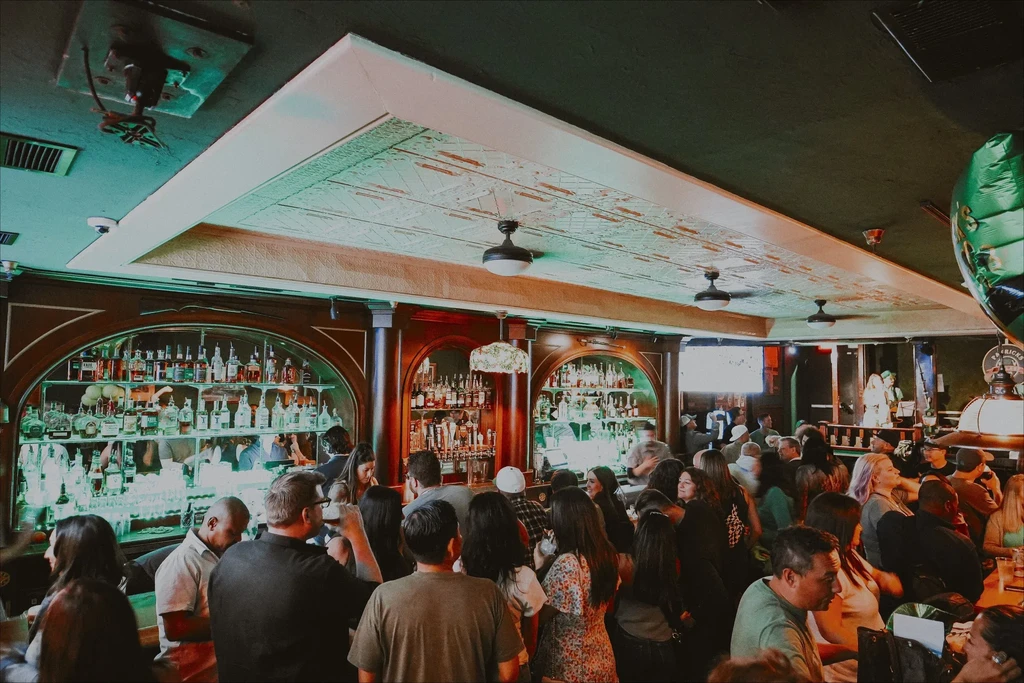 Crowded pub interior shows ornate bar, green lit bottles, and St. Patrick's sign.