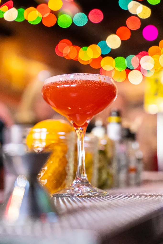 Red frothy cocktail in coupe glass, blurred colorful bar lights.