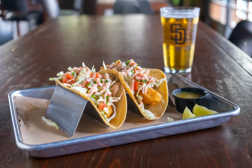 Two fried fish tacos with lime, green sauce, and beer.
